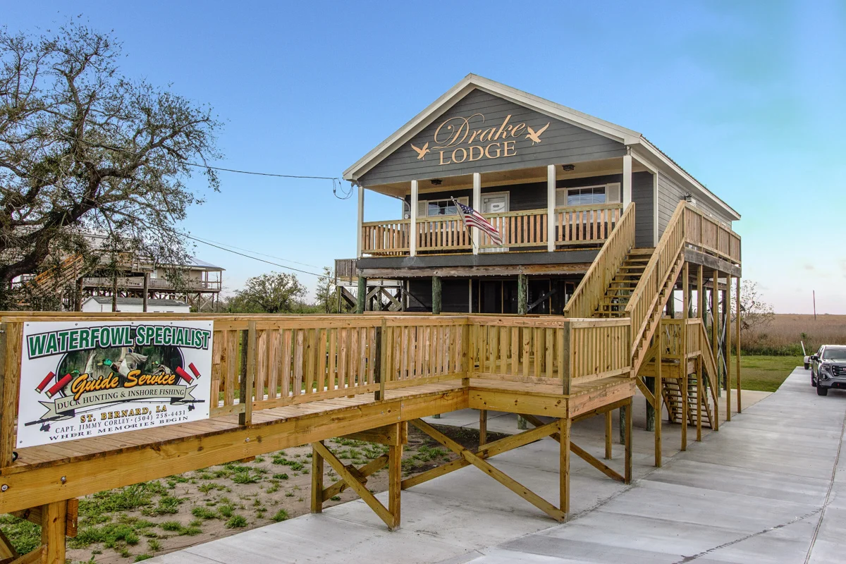 Drake Lodge exterior with wooden ramp, Waterfowl Specialist guide service sign, American flag, and marshland backdrop in St. Bernard, Louisiana.
