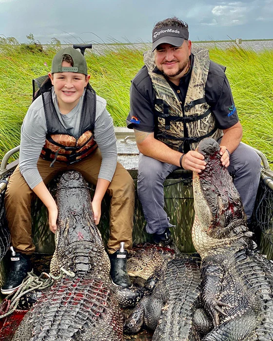 Two hunters, a boy and a man, posing with large alligators in a boat surrounded by tall grass, showcasing a successful alligator hunt in Louisiana's wetlands.