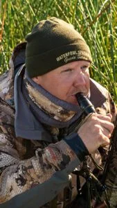 Man in camouflage gear holding a duck call in a marsh setting, representing Waterfowl Specialist Guide Service's duck hunting expertise in Louisiana.